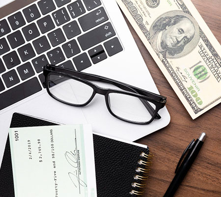 Laptop, 100 dollar bill, glasses, check and pen on a table