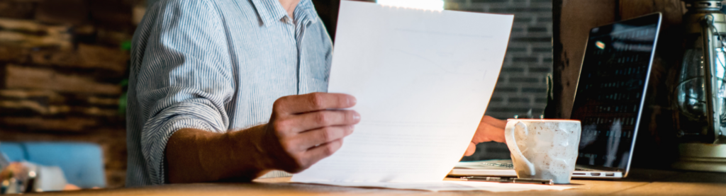 Close up image of man holding paper with laptop and coffee cup in background.