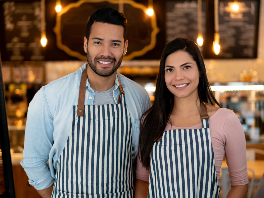 Male and female cafe managers wearing stripped aprons smiling at camera.